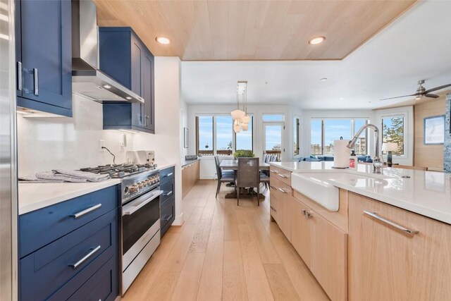 kitchen featuring wall chimney range hood, stainless steel stove, sink, blue cabinetry, and hanging light fixtures