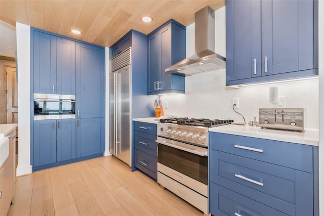 kitchen featuring wall chimney exhaust hood, blue cabinets, light hardwood / wood-style flooring, wooden ceiling, and appliances with stainless steel finishes