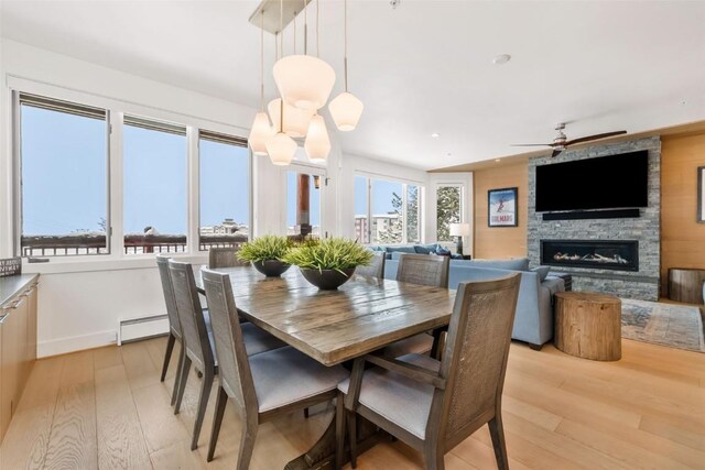 dining room with a baseboard heating unit, a stone fireplace, ceiling fan with notable chandelier, and a healthy amount of sunlight