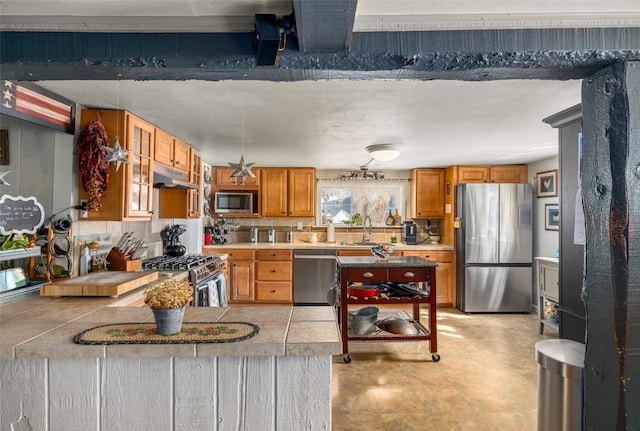 kitchen with brown cabinetry, a peninsula, stainless steel appliances, under cabinet range hood, and a sink