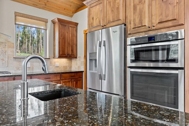 kitchen featuring backsplash, appliances with stainless steel finishes, brown cabinets, a sink, and dark stone counters