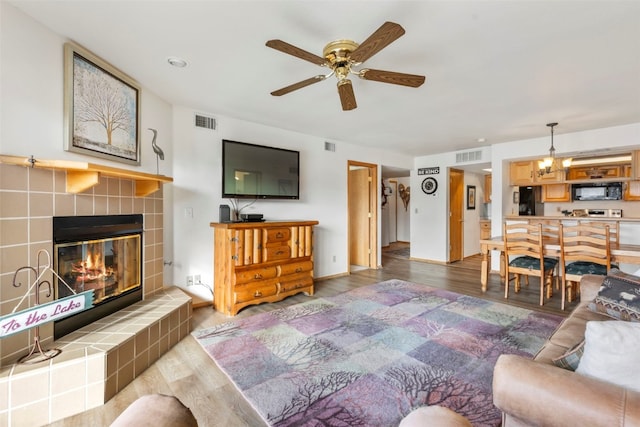 living area featuring ceiling fan with notable chandelier, a tiled fireplace, wood finished floors, and visible vents