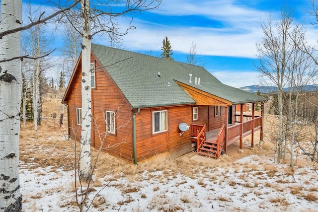 snow covered back of property with a shingled roof and a deck