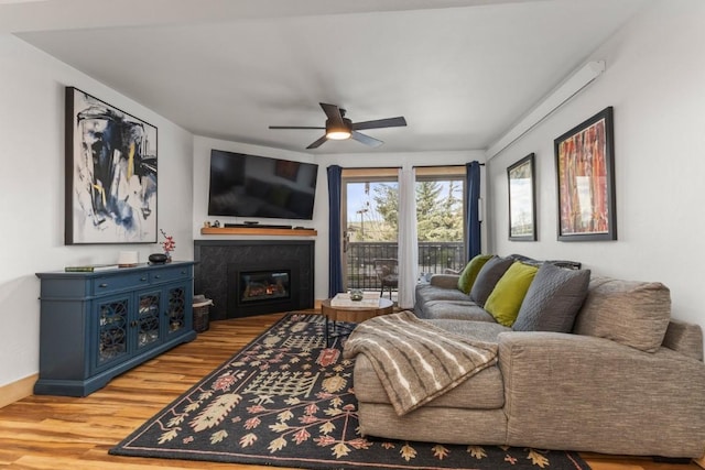 living area with wood finished floors, a glass covered fireplace, a ceiling fan, and baseboards