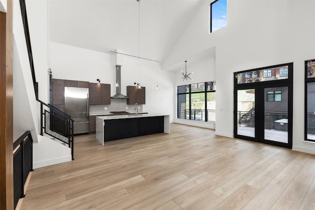 kitchen with an island with sink, hanging light fixtures, stainless steel appliances, dark brown cabinets, and wall chimney range hood