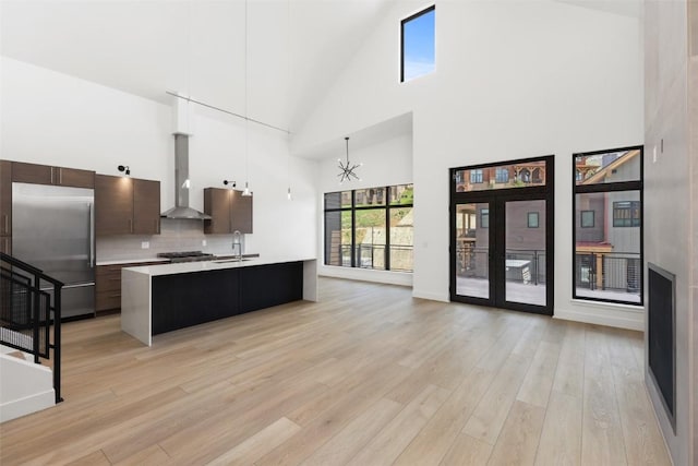 kitchen with dark brown cabinets, appliances with stainless steel finishes, pendant lighting, a kitchen island with sink, and wall chimney range hood