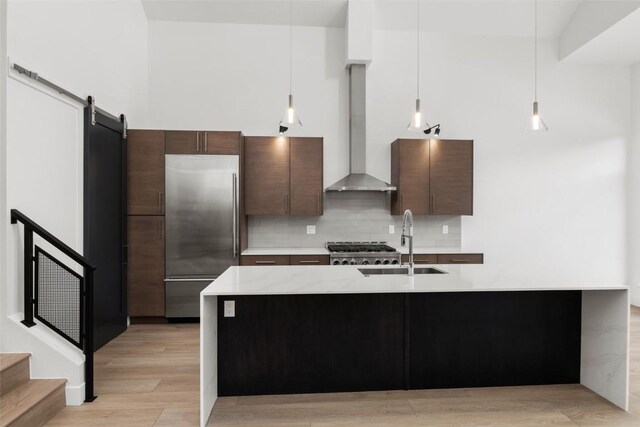 kitchen featuring dark brown cabinets, wall chimney range hood, a barn door, decorative light fixtures, and stainless steel built in fridge