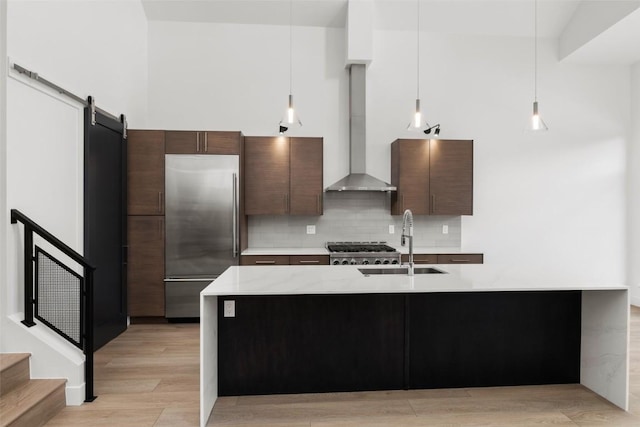 kitchen featuring wall chimney exhaust hood, decorative light fixtures, dark brown cabinets, built in fridge, and a barn door