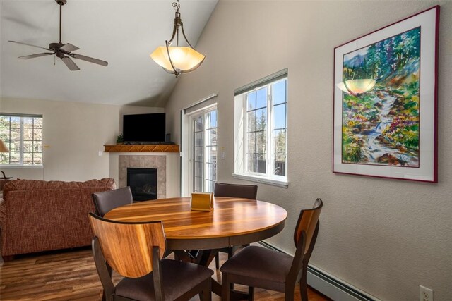 dining room with a baseboard heating unit, a tiled fireplace, ceiling fan, high vaulted ceiling, and dark wood-type flooring