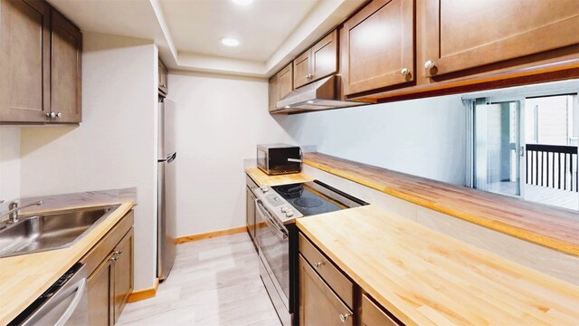 kitchen with butcher block countertops, a sink, stainless steel appliances, baseboards, and under cabinet range hood