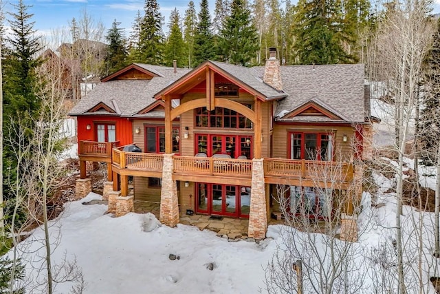 snow covered house featuring a wooden deck, stone siding, roof with shingles, french doors, and a chimney