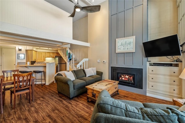 living area featuring a high ceiling, ceiling fan, stairway, a fireplace, and dark wood-type flooring