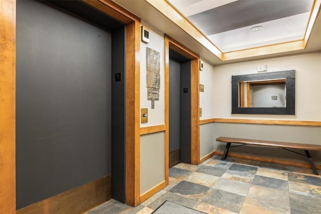 hallway featuring a tray ceiling, elevator, stone finish flooring, and baseboards