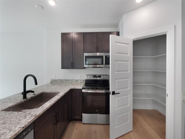 kitchen with stainless steel appliances, a sink, dark brown cabinets, light wood finished floors, and light stone counters