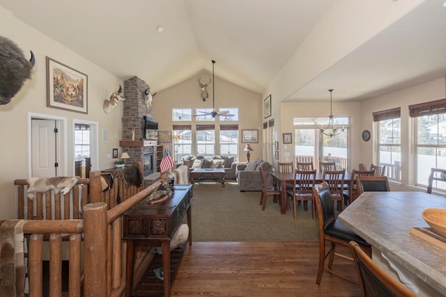 dining space featuring ceiling fan with notable chandelier, dark wood-style floors, a large fireplace, and high vaulted ceiling