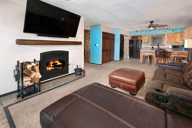 living area with light carpet, a warm lit fireplace, rail lighting, and ceiling fan