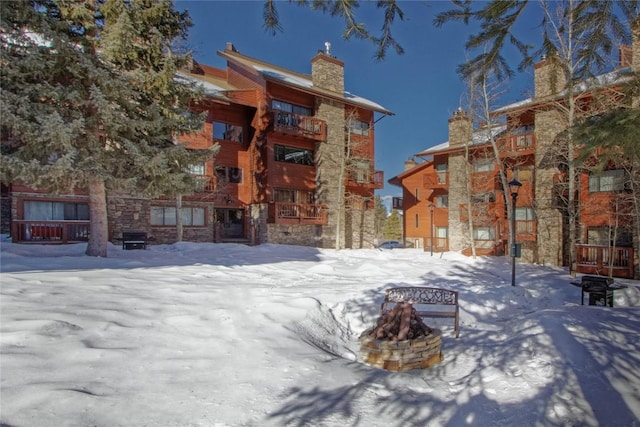 snow covered rear of property featuring stone siding, an outdoor fire pit, and a chimney