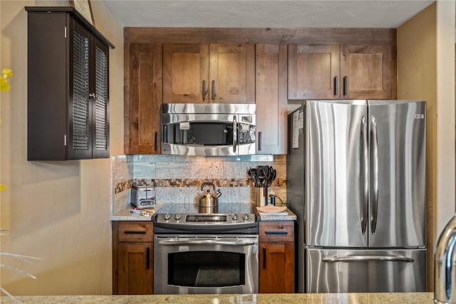 kitchen with brown cabinetry, decorative backsplash, light stone counters, and stainless steel appliances