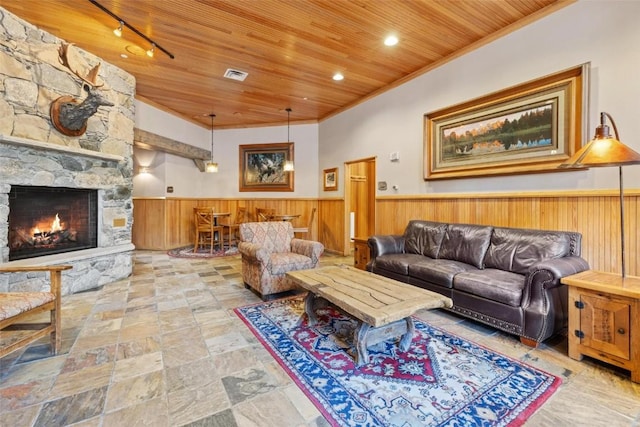 living room featuring wainscoting, wood ceiling, wooden walls, a stone fireplace, and ornamental molding