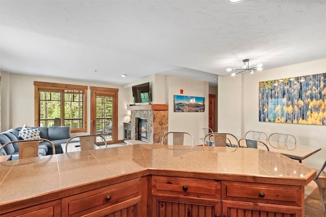 kitchen with tile counters, a stone fireplace, brown cabinets, open floor plan, and a chandelier