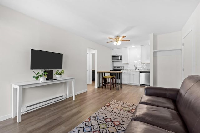 living area with baseboard heating, light wood finished floors, a ceiling fan, and baseboards