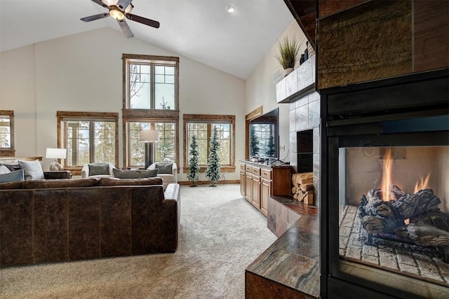 living area featuring baseboards, a ceiling fan, light colored carpet, a lit fireplace, and high vaulted ceiling