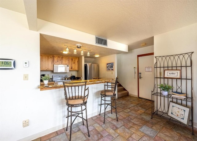 kitchen featuring visible vents, a peninsula, white appliances, a kitchen breakfast bar, and stone finish floor