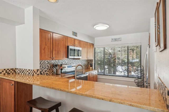 kitchen with brown cabinets, appliances with stainless steel finishes, a peninsula, decorative backsplash, and light stone countertops