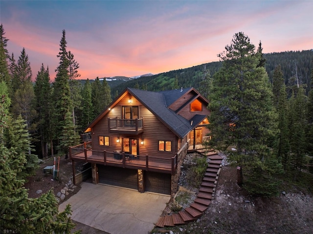 back of property with a forest view, a balcony, driveway, an attached garage, and a mountain view