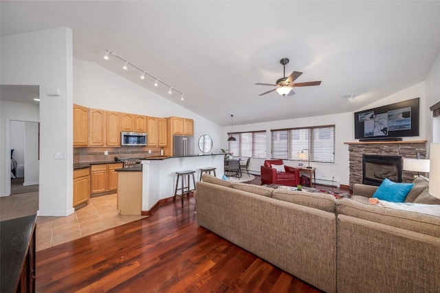 living room featuring vaulted ceiling, a stone fireplace, light wood-type flooring, a baseboard radiator, and ceiling fan