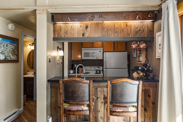 kitchen featuring dark countertops, a baseboard heating unit, brown cabinets, white microwave, and freestanding refrigerator