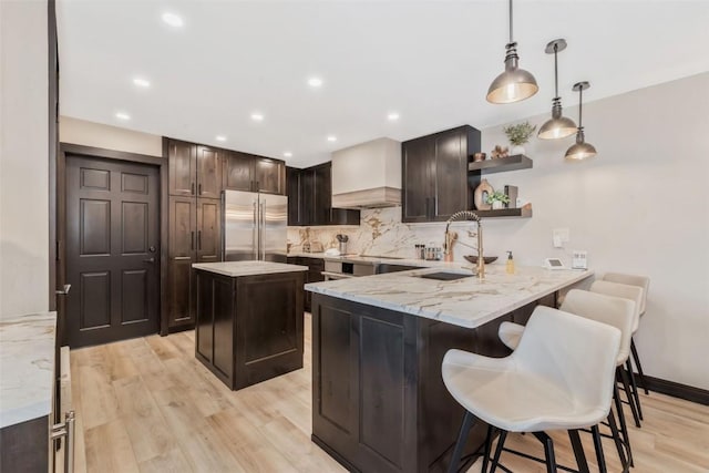 kitchen with light wood finished floors, open shelves, custom range hood, dark brown cabinets, and built in refrigerator