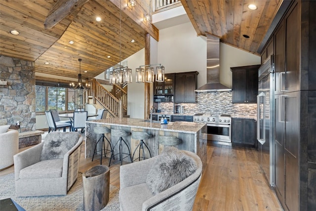 kitchen with light stone countertops, stainless steel appliances, wall chimney range hood, backsplash, and a kitchen island with sink