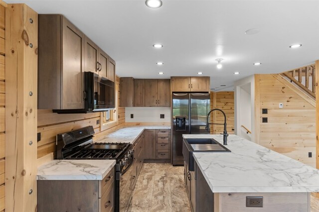 kitchen with light stone counters, sink, black appliances, wood walls, and an island with sink