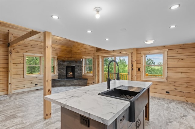 kitchen featuring a stone fireplace, wood walls, an island with sink, and light stone counters