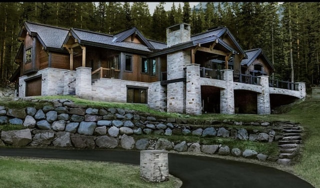 back of property featuring a balcony, a forest view, a chimney, a garage, and stone siding