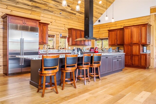 kitchen featuring light stone countertops, island exhaust hood, high vaulted ceiling, stainless steel built in fridge, and wood walls