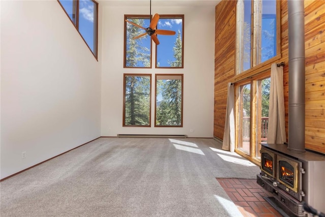 unfurnished living room with a wood stove, ceiling fan, a high ceiling, a baseboard heating unit, and dark carpet