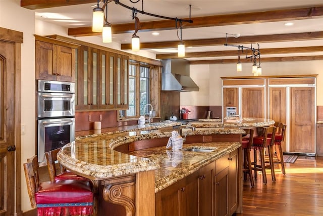 kitchen featuring a large island, wall chimney exhaust hood, decorative light fixtures, beam ceiling, and stainless steel double oven