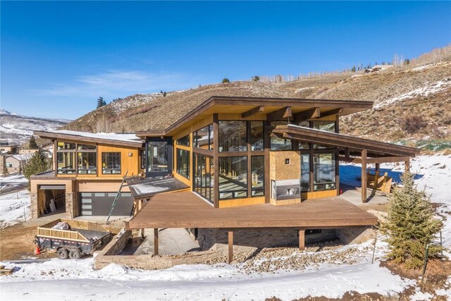 snow covered rear of property featuring a garage, a mountain view, and a sunroom