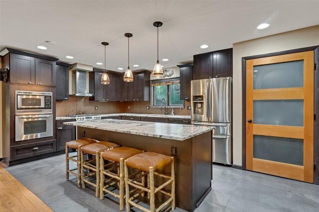 kitchen featuring wall chimney exhaust hood, dark brown cabinets, stainless steel appliances, pendant lighting, and a center island