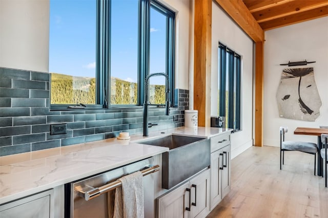 kitchen featuring light wood-type flooring, backsplash, stainless steel dishwasher, light stone counters, and beamed ceiling