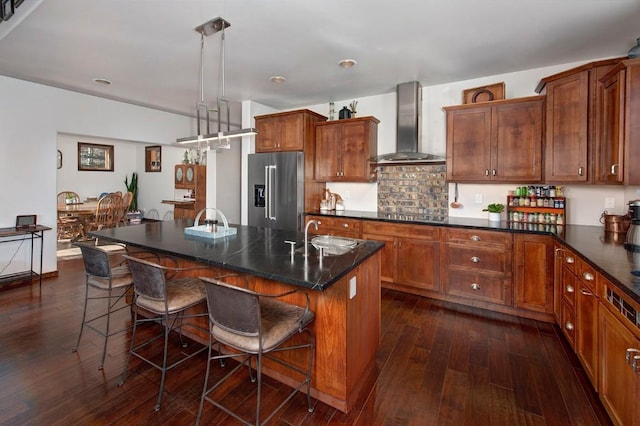 kitchen with high end fridge, sink, wall chimney range hood, decorative light fixtures, and an island with sink