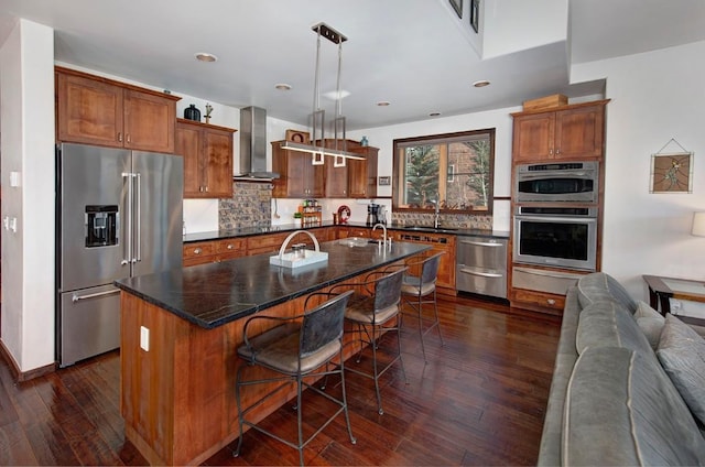 kitchen with appliances with stainless steel finishes, wall chimney exhaust hood, a breakfast bar, hanging light fixtures, and an island with sink