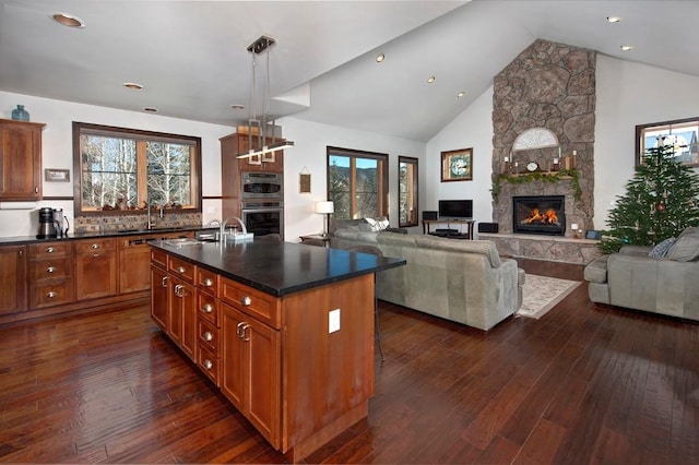 kitchen featuring backsplash, a stone fireplace, an island with sink, decorative light fixtures, and stainless steel double oven