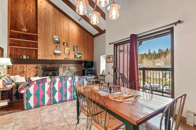 dining space featuring a baseboard heating unit, high vaulted ceiling, tile patterned flooring, a stone fireplace, and wood walls