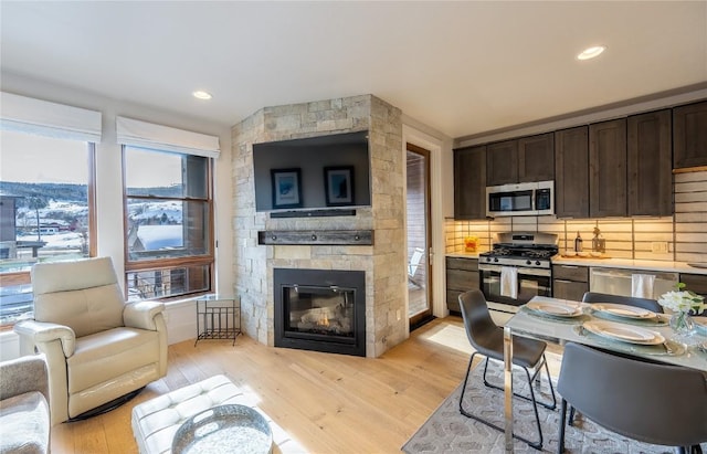 kitchen with stainless steel appliances, dark brown cabinetry, tasteful backsplash, a stone fireplace, and light wood-type flooring