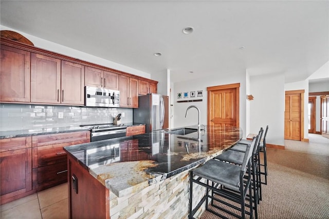 kitchen with a kitchen island with sink, a sink, appliances with stainless steel finishes, tasteful backsplash, and dark stone countertops