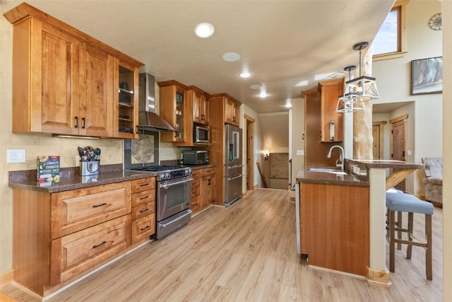 kitchen featuring light wood-type flooring, appliances with stainless steel finishes, a peninsula, a kitchen breakfast bar, and wall chimney exhaust hood