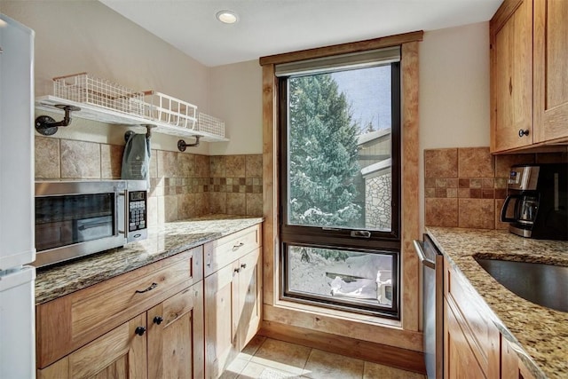 kitchen with decorative backsplash, appliances with stainless steel finishes, brown cabinetry, light tile patterned flooring, and light stone countertops